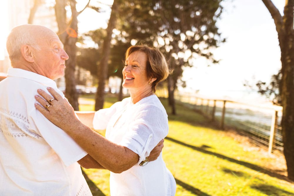 Older couple dressed all in white smiles and dances together outdoors Charter Senior Living Residents couple dressed all in white smiles and dances together outdoors