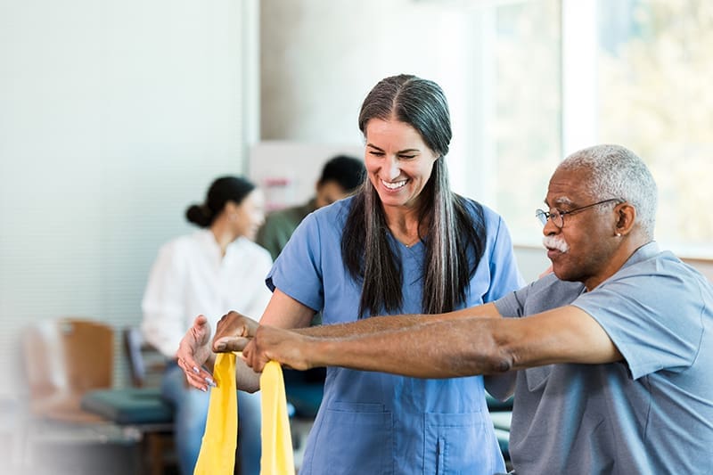 As other therapists work with patients in the background, the mid adult female physical therapist teaches the senior adult man how to use an elastic band to exercise his arms.